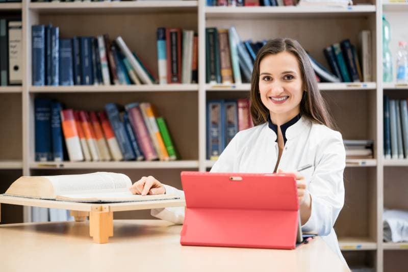 Medical student in library researching books and writing down results on her laptop computer